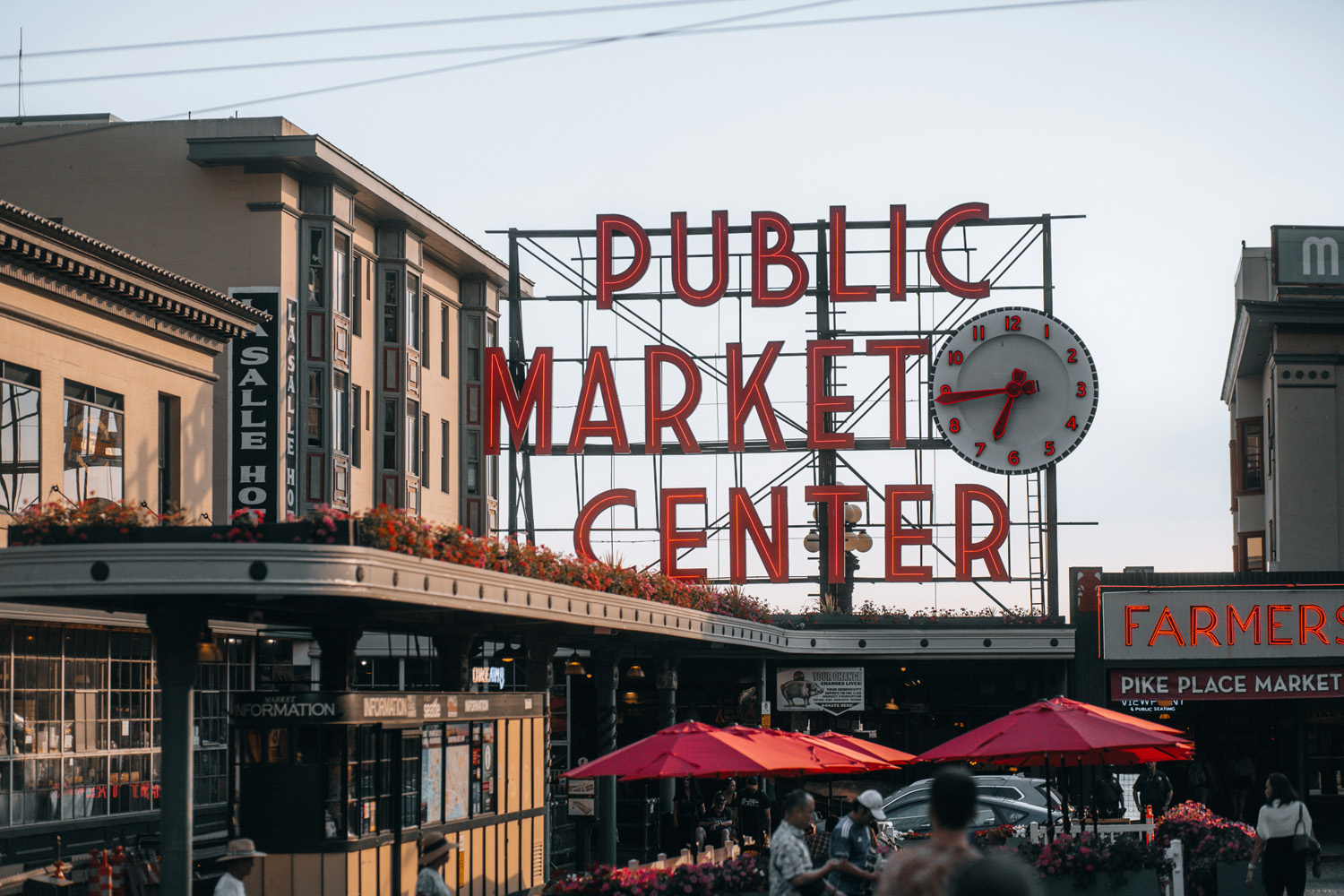 The iconic red neon sign of Public Market Center at Pike Place Market in Seattle, with flower boxes and crowds beneath red umbrellas.