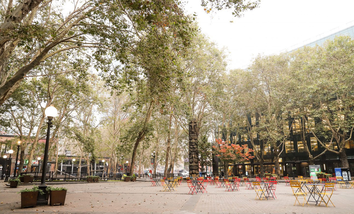 Tree-lined open plaza at Occidental Square in Seattle, featuring colorful outdoor seating, lamp posts, and public art installations.