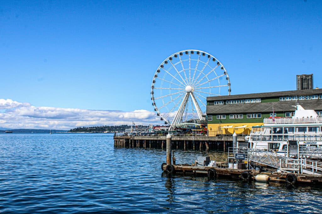 Bright blue waterfront scene in Seattle showing the Seattle Great Wheel, tour boats, and part of the pier against a sunny sky.