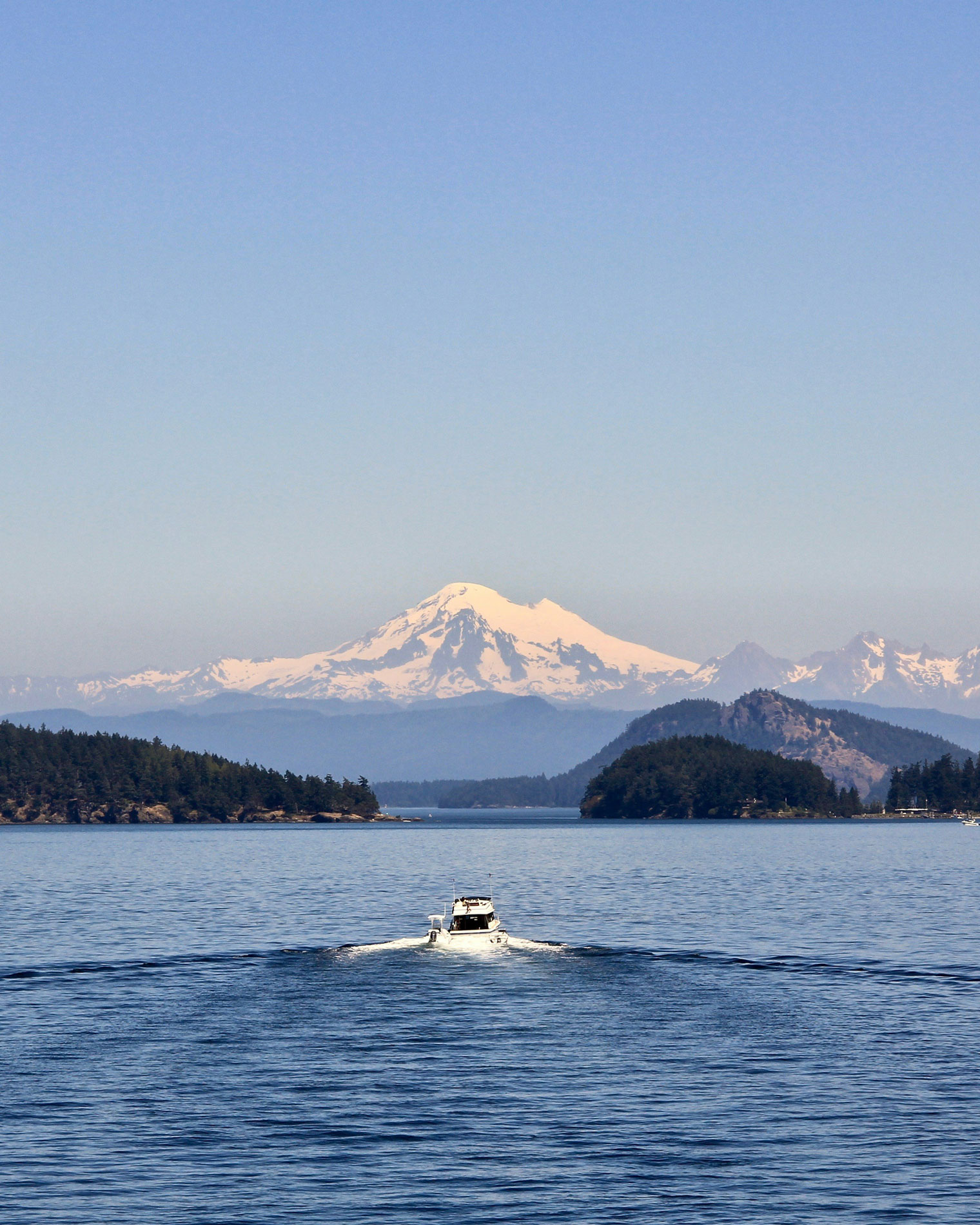 A white motorboat leaving a gentle wake as it cruises through calm blue waters, surrounded by forested islands, with snow-covered Mount Baker and the Cascade Range rising in the background under a clear sky.