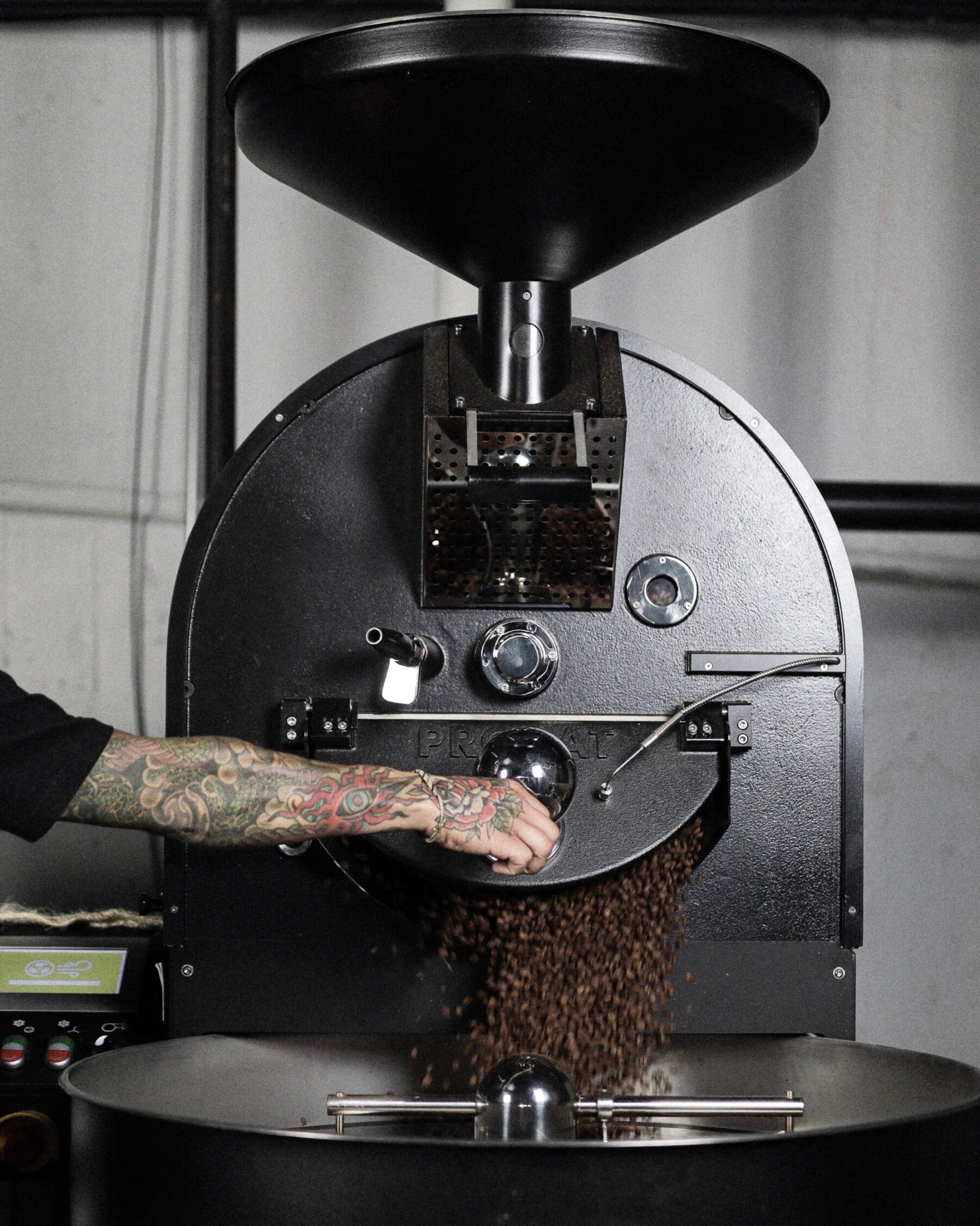 Tattooed barista’s hand operating a commercial coffee roaster as freshly roasted beans pour out into a cooling tray, with a matte black Probat machine as the backdrop in a minimal industrial space.