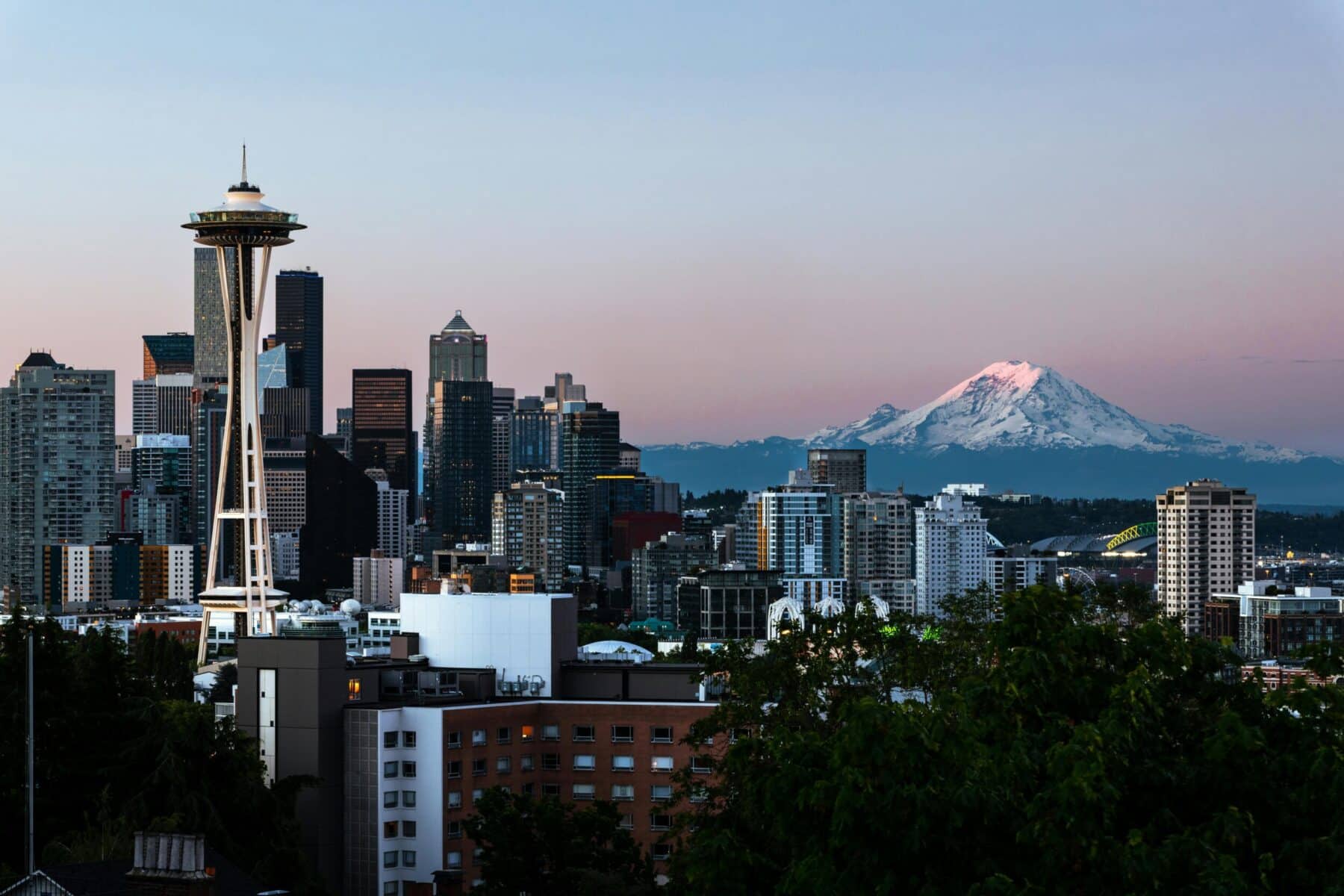 Panoramic view of downtown Seattle at dusk, featuring the Space Needle in the foreground, modern skyscrapers throughout the cityscape, and snow-capped Mount Rainier glowing in the distance under a pink and blue sky.