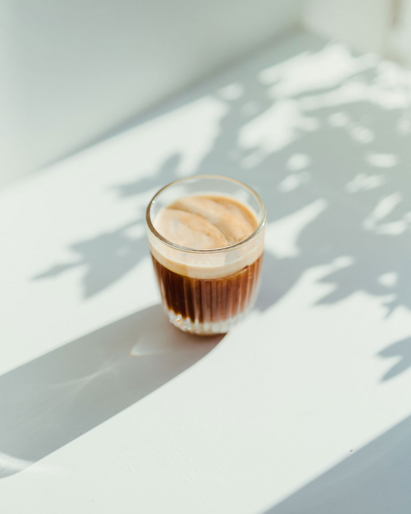 A glass of espresso with a creamy top layer sitting on a white surface, surrounded by soft shadows of leaves cast by sunlight streaming through a nearby window.