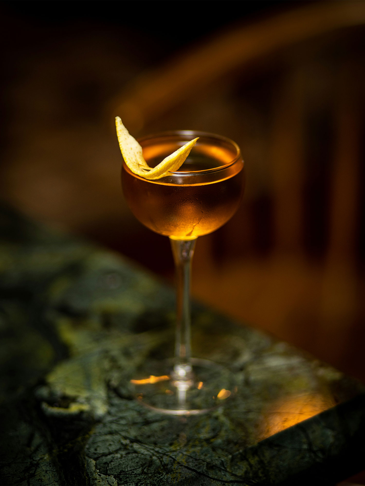 A close-up of a chilled amber cocktail garnished with a lemon twist, served in a coupe glass on a green marble bar, with warm ambient lighting in the background.