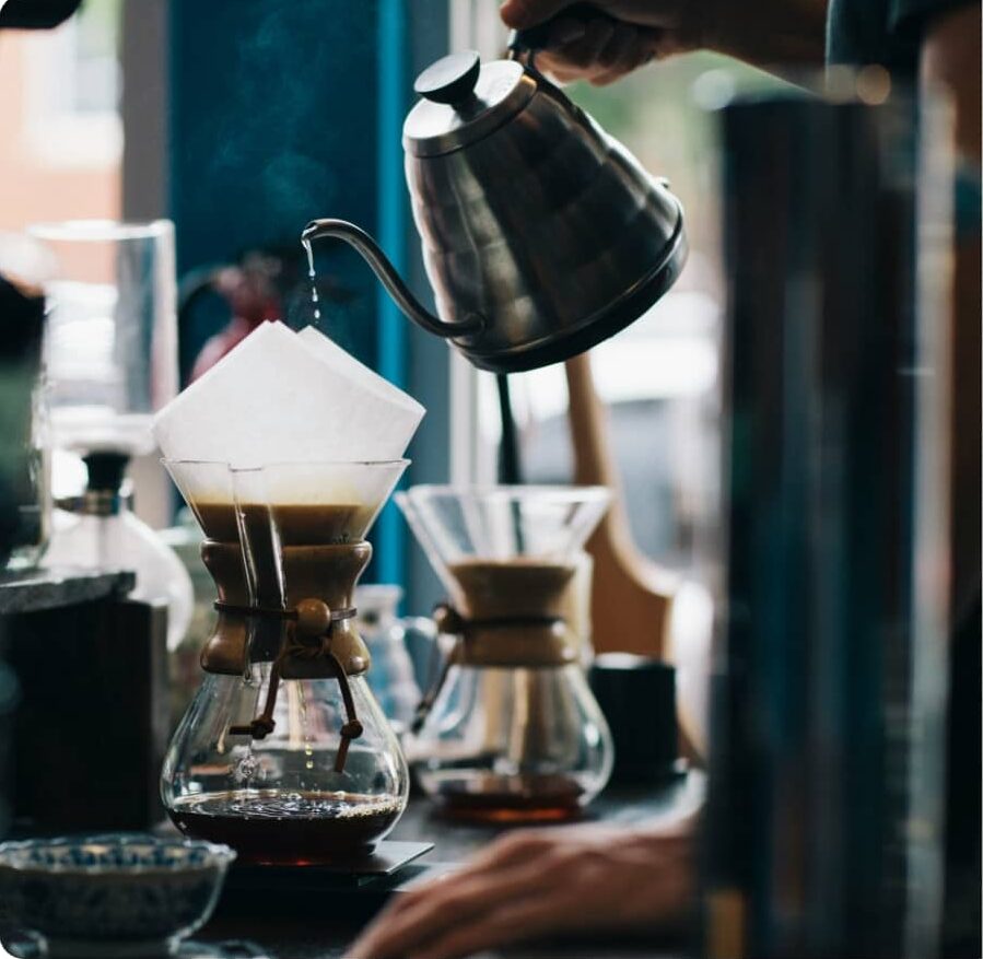 Hand pouring hot water from a gooseneck kettle into a paper filter atop a Chemex coffee maker, with freshly brewed coffee collecting below in a warm, softly lit café setting.