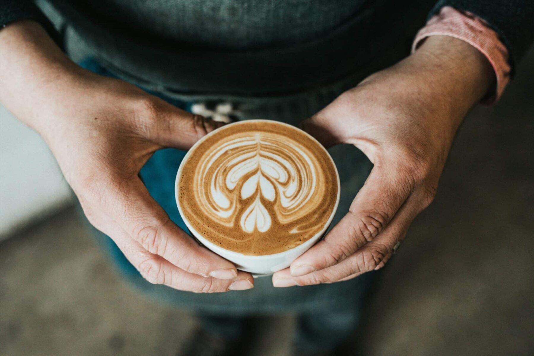 Close-up of a person holding a cup of latte with intricate heart-shaped foam art, framed by both hands and set against a blurred background and dark denim apron.