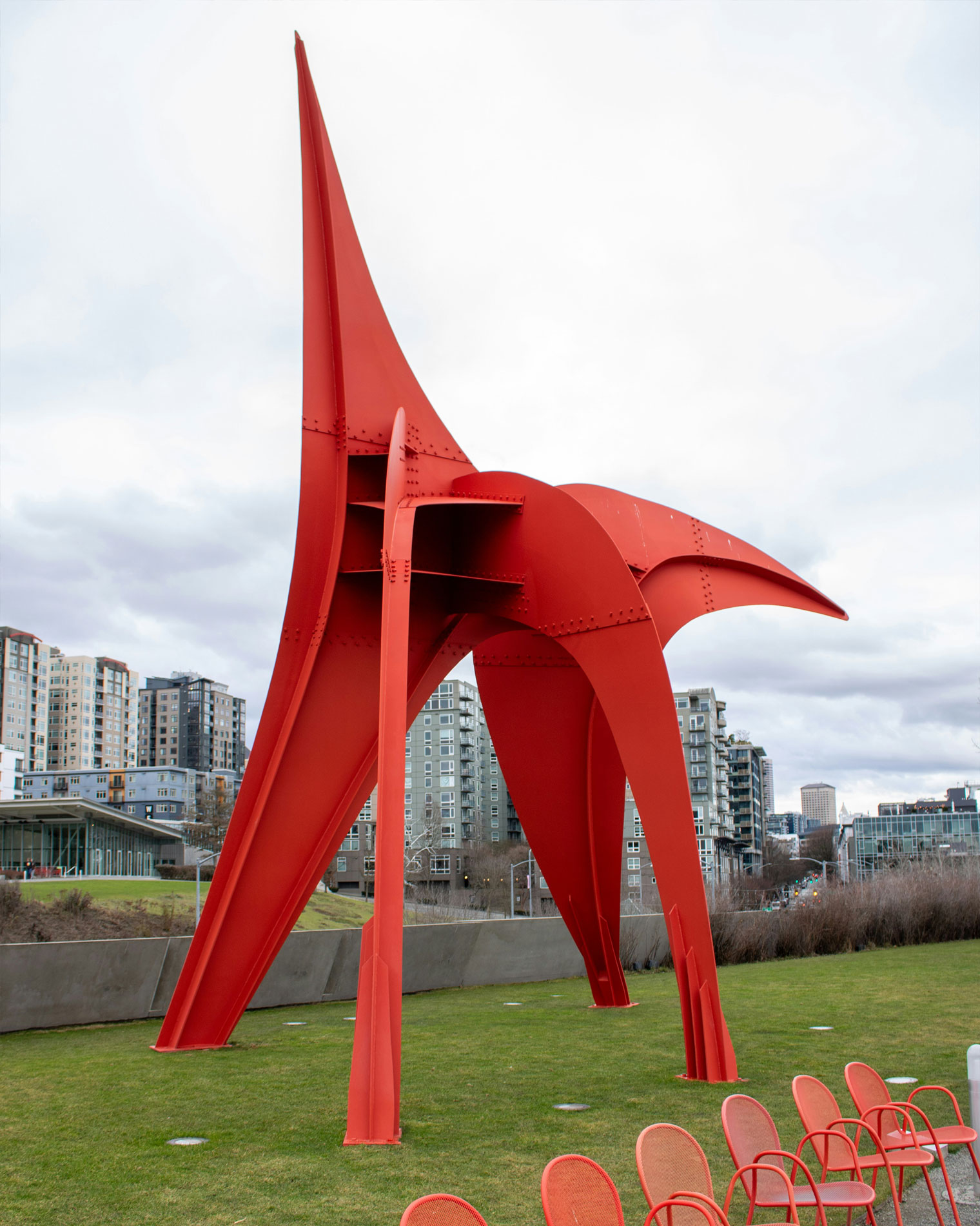 Large red steel sculpture titled "Eagle" by Alexander Calder, displayed on a grassy lawn at Seattle’s Olympic Sculpture Park, with modern city buildings in the background under a cloudy sky.