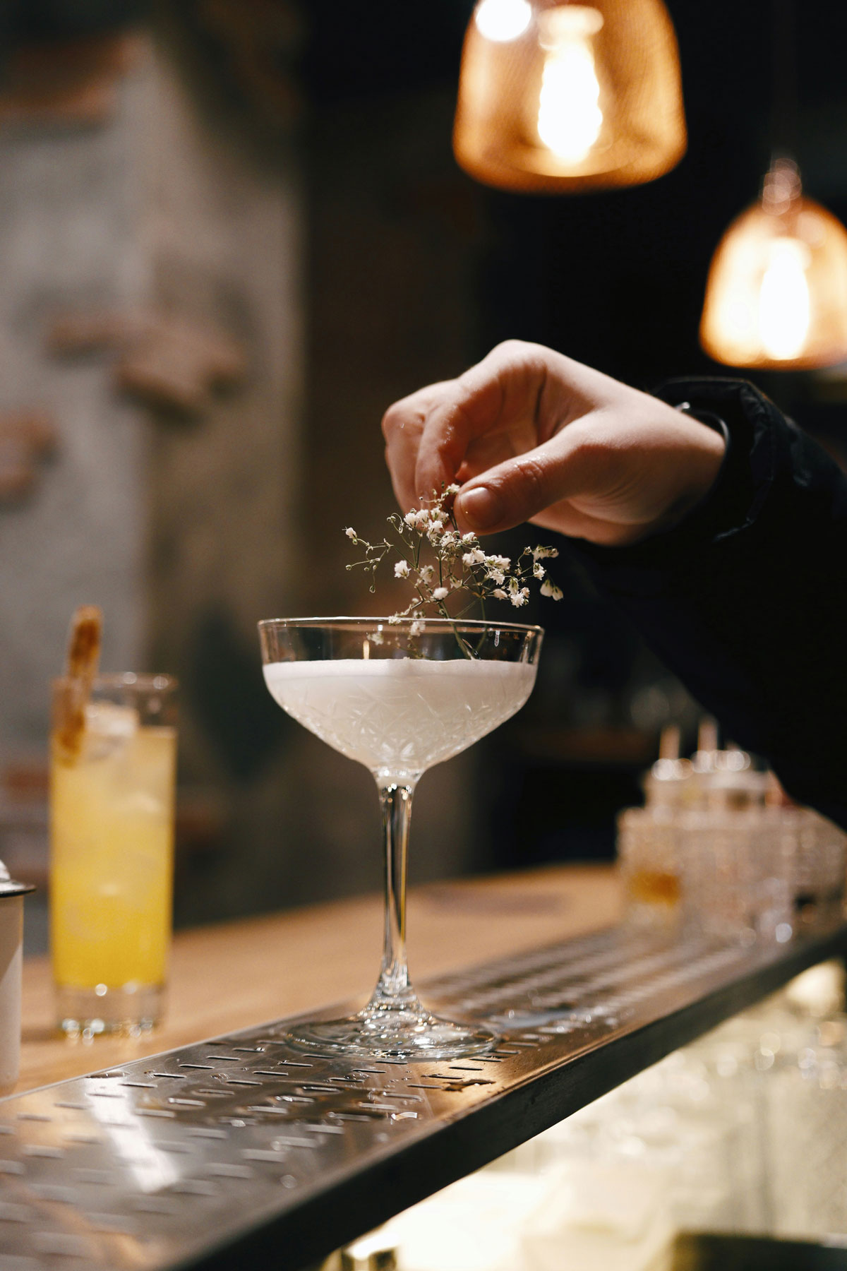 A hand delicately places a small floral garnish on a cocktail in a crystal glass. The setting is a cozy bar with warm, ambient lighting from hanging lamps. Another cocktail with a bright yellow hue and a straw is slightly blurred in the background. The metallic bar surface reflects the soft, golden glow of the surroundings. The image is framed to focus on the artistic touch of cocktail preparation, conveying a sense of elegance and craftsmanship.