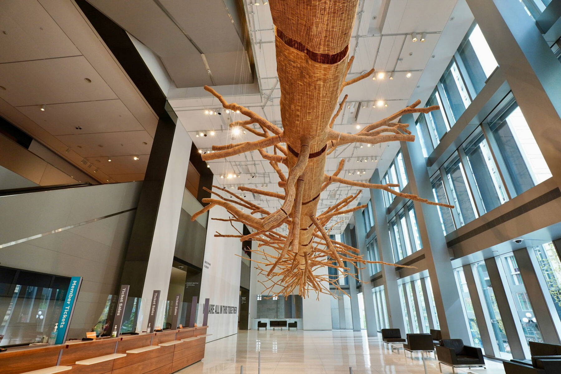 Large woven tree sculpture suspended from the ceiling of the Seattle Art Museum lobby, surrounded by modern architecture with tall glass windows, ticket counters, and polished floors.