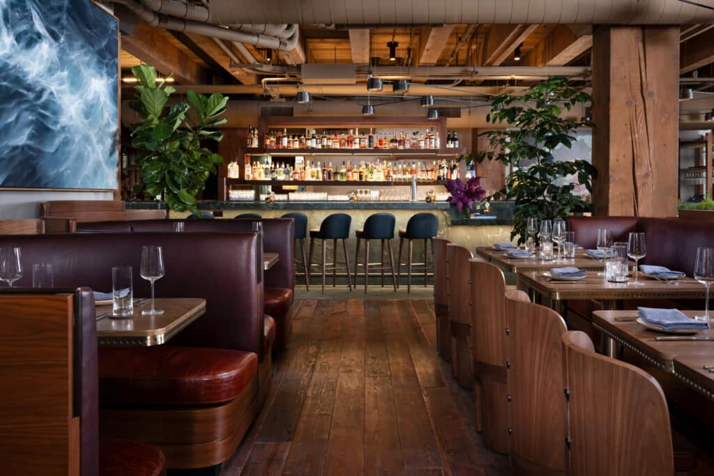 Interior of Salt Harvest restaurant, showing leather booth seating and a wooden bar with hanging string lights, exposed wood beams, and potted plants in a warm, rustic dining room