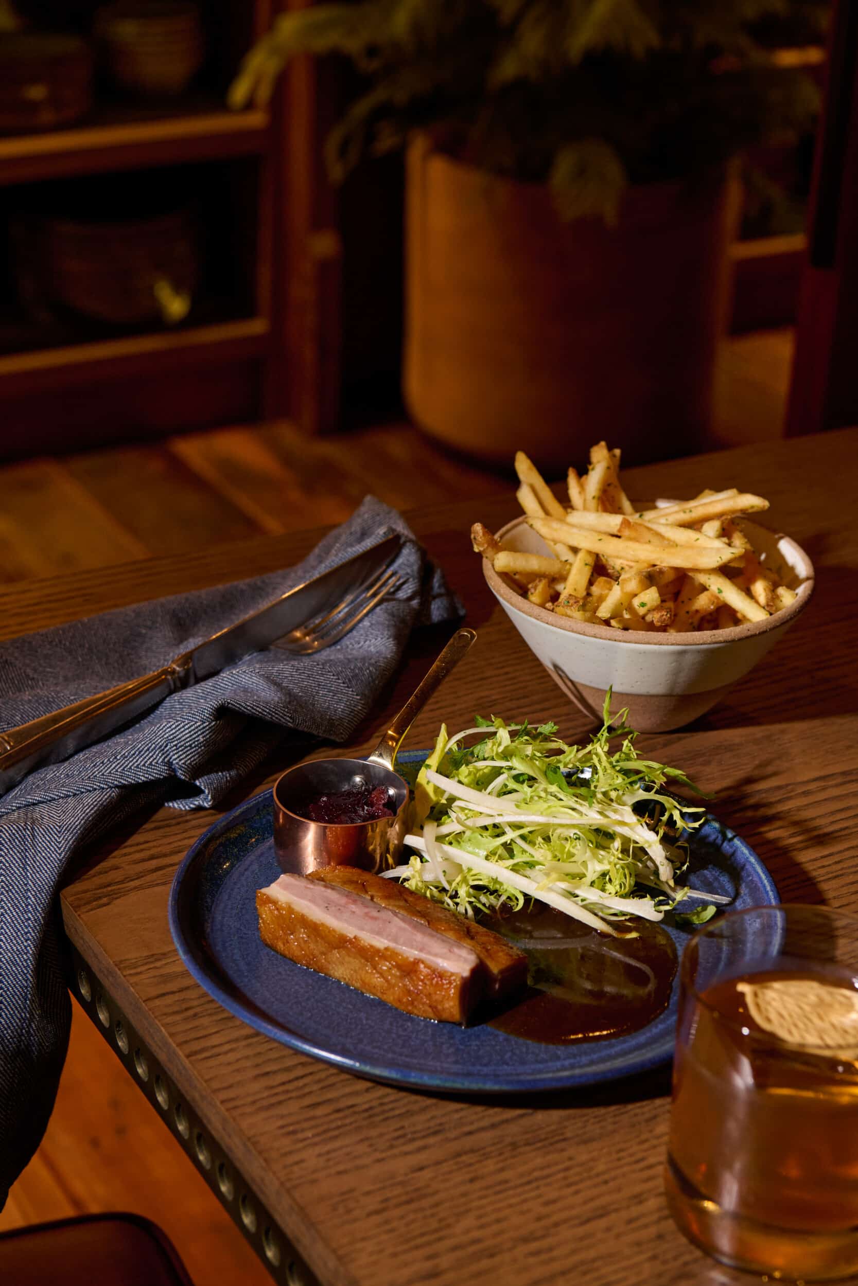 Plated dish at Salt Harvest featuring a glazed meat entrée with shredded cabbage slaw and a small pitcher of sauce, served alongside a bowl of shoestring fries on a wooden table